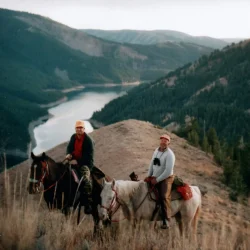 Two people on horseback overlook a winding river and forested mountains under a cloudy sky.