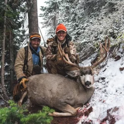 Two hunters pose in snowy woods with a large deer they have hunted, both wearing orange hats and winter gear.