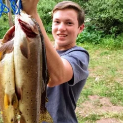 Teenager holding up three freshly caught fish on a blue stringer, standing outdoors with trees and grass in the background.