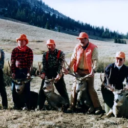 Five hunters in outdoor gear kneel behind four deer with large antlers on a grassy, mountainous landscape.