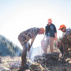Three people in camouflage and orange hats gather around a small campfire in a forested mountain area during daylight.