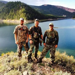 Three men in camouflage outfits stand on a rocky hill overlooking a lake surrounded by forested mountains.