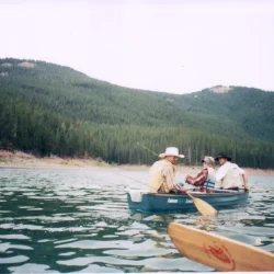 Three people sit in a boat on a lake, fishing, with forested hills in the background and a paddle in the foreground.