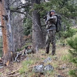 Two people in camouflage gear, one standing and one lying down, hunting or scouting in a wooded forest area.