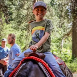 A smiling child in a cap and green shirt rides a horse in a wooded area, with two adults in the background.