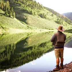 Person in hat and boots fishes at the edge of a calm lake with green hills reflected in the water.