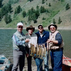 Four men pose by a lake with fishing gear, holding a line of six caught fish in front of canoes and a forested hillside.