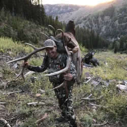 Person in camouflage carries elk antlers and hide on their back in a mountainous, forested landscape at sunset.