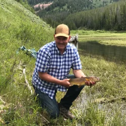Man in plaid shirt and cap kneels by a grassy riverbank holding a fish with forested hills in the background.