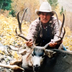Person in a cowboy hat poses outdoors, holding the antlers of a large deer lying on the ground.