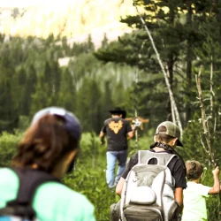 Three people with backpacks hike on a forest trail, following a person wearing a cowboy hat ahead of them.