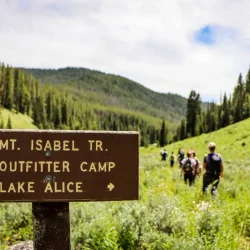 Trail sign pointing to Mt. Isabel Tr., Outfitter Camp, and Lake Alice; hikers walk on a grassy trail in background.