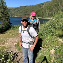 Man carrying a child in a hiking backpack near a lake, surrounded by trees and mountains on a sunny day.