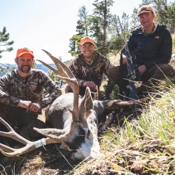 Three hunters in camouflage and orange hats pose with a large deer they hunted in a grassy, mountainous area.